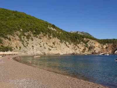 A small-pebble beach with calm water with a green hill in the background and a small boat on the right