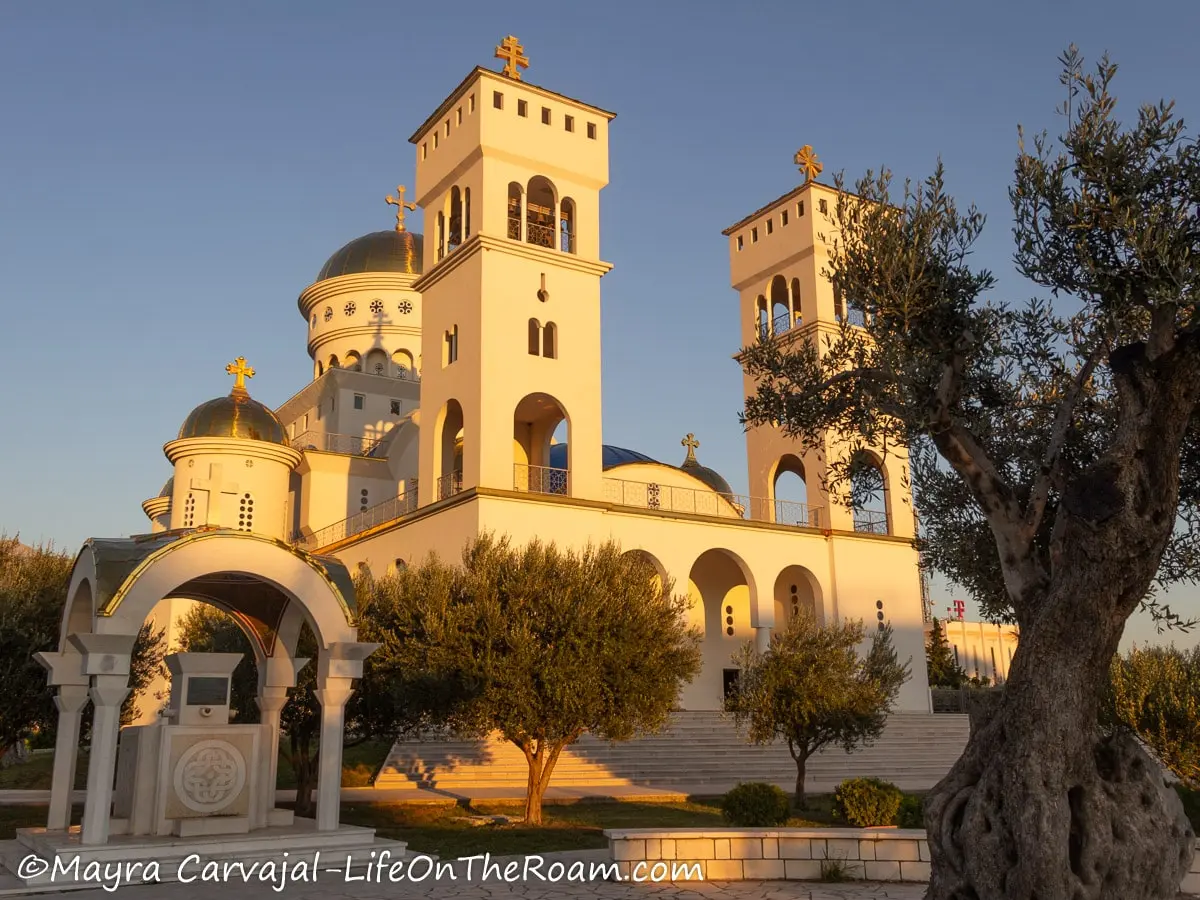 A large church in white with tall towers on each side and golden domes, with and olive garden in front