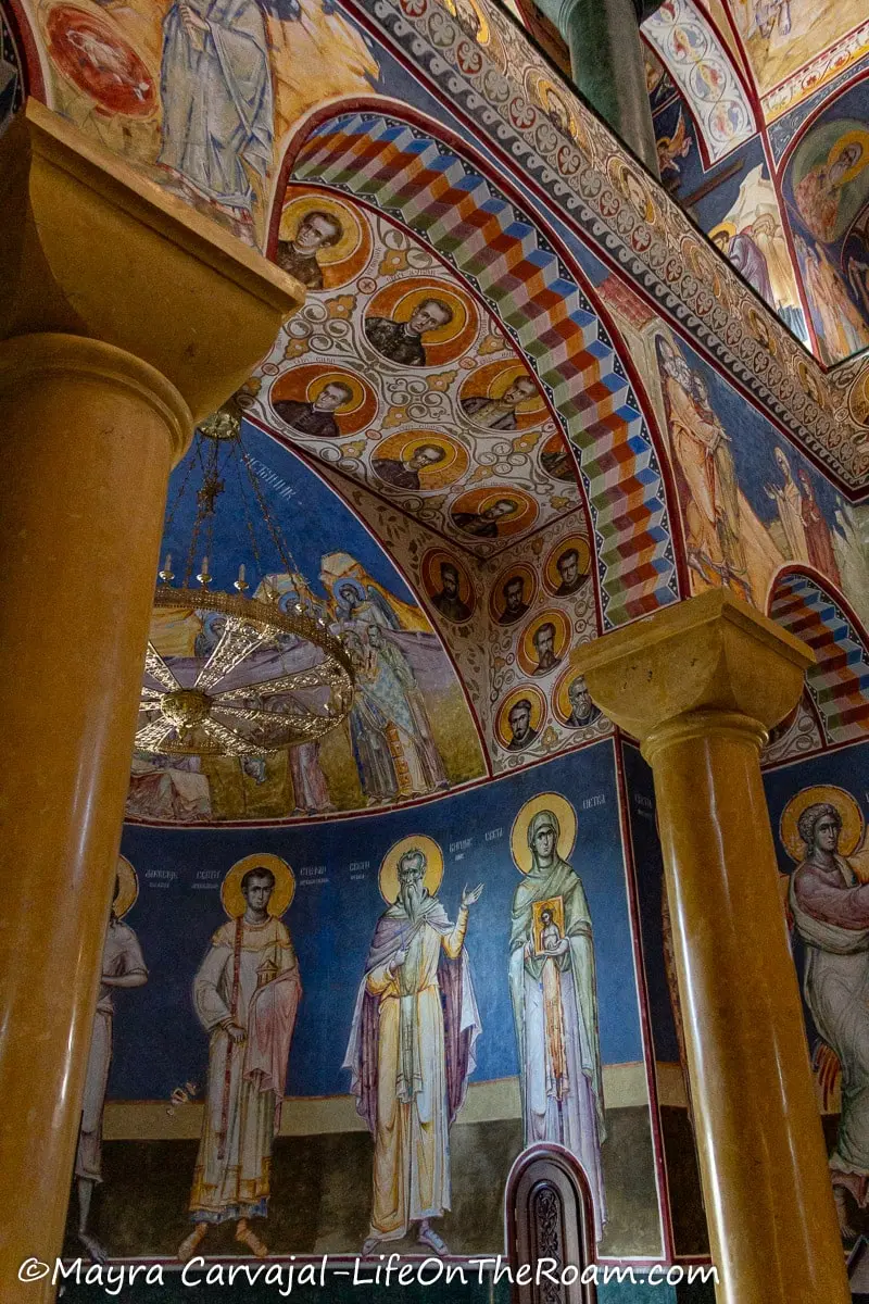 A trompe l'oeil decorating the underside of an arch ending in two stone columns, inside a church with tall ceilings with walls covered in murals depicting saints