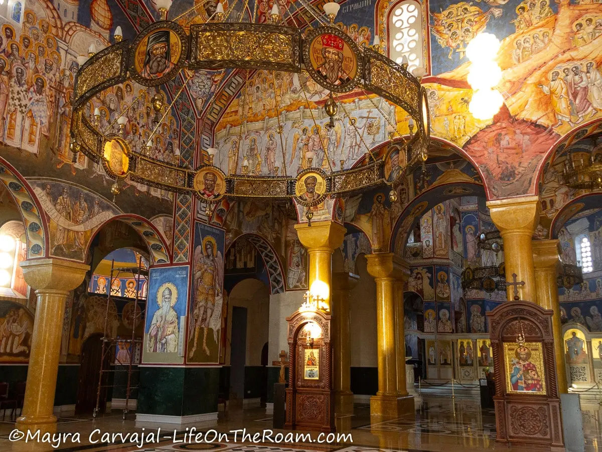 A round huge, decorated, metal gold plated light fixture hanging in a church with walls covered in murals