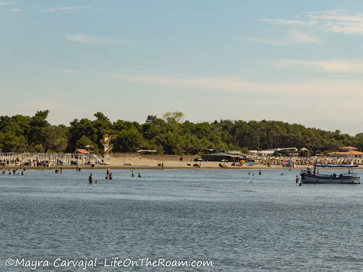 View in the distance of a long, busy, sandy beach with boats on the water and trees at the back