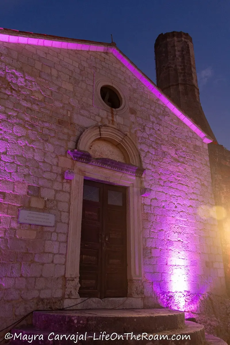 An old stone building with a double door in wood with an arch on top, illuminated with a purple light