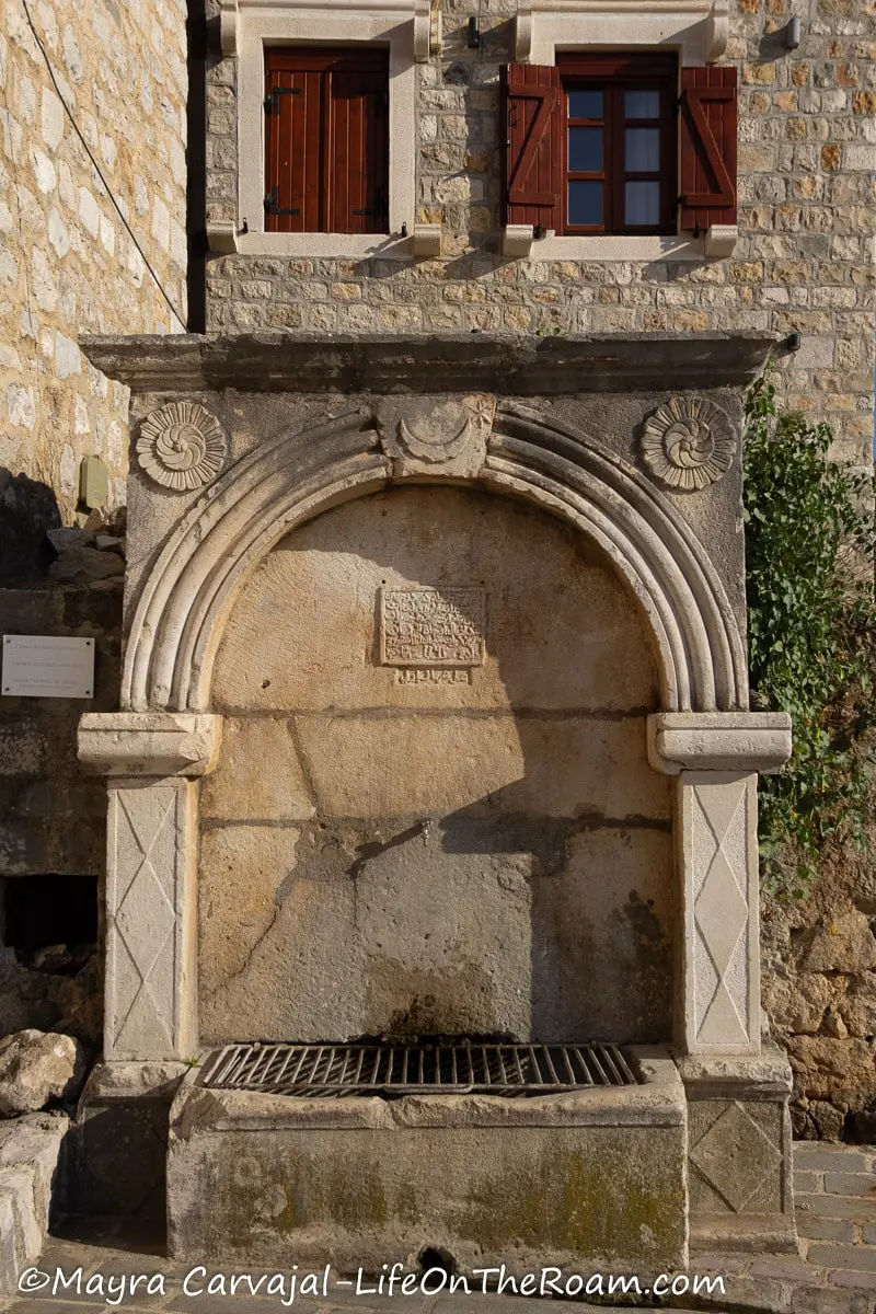 A water fountain in stone with an arch supported by columns, with floral motifs on the upper corners