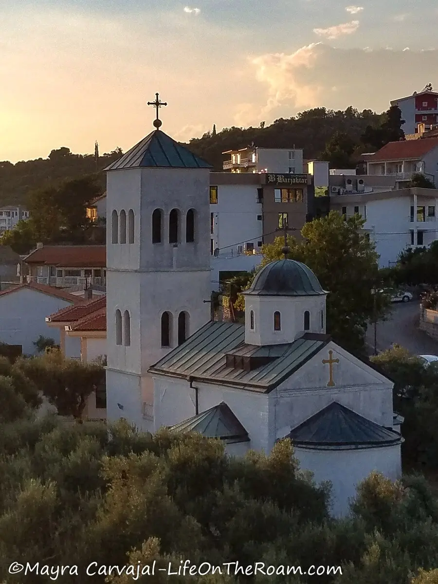 View of a small church from above with a tower topped with a cross and a small dome on the side, surrounded by olive trees