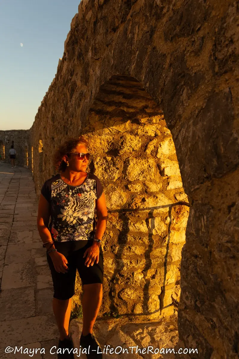 Mayra standing next to an arched opening on a fortress ancient wall looking at the sunset