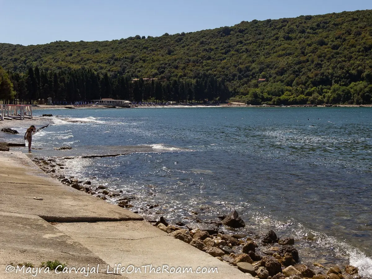 A broken concrete path alongside a beach with tree-covered hills in the distance