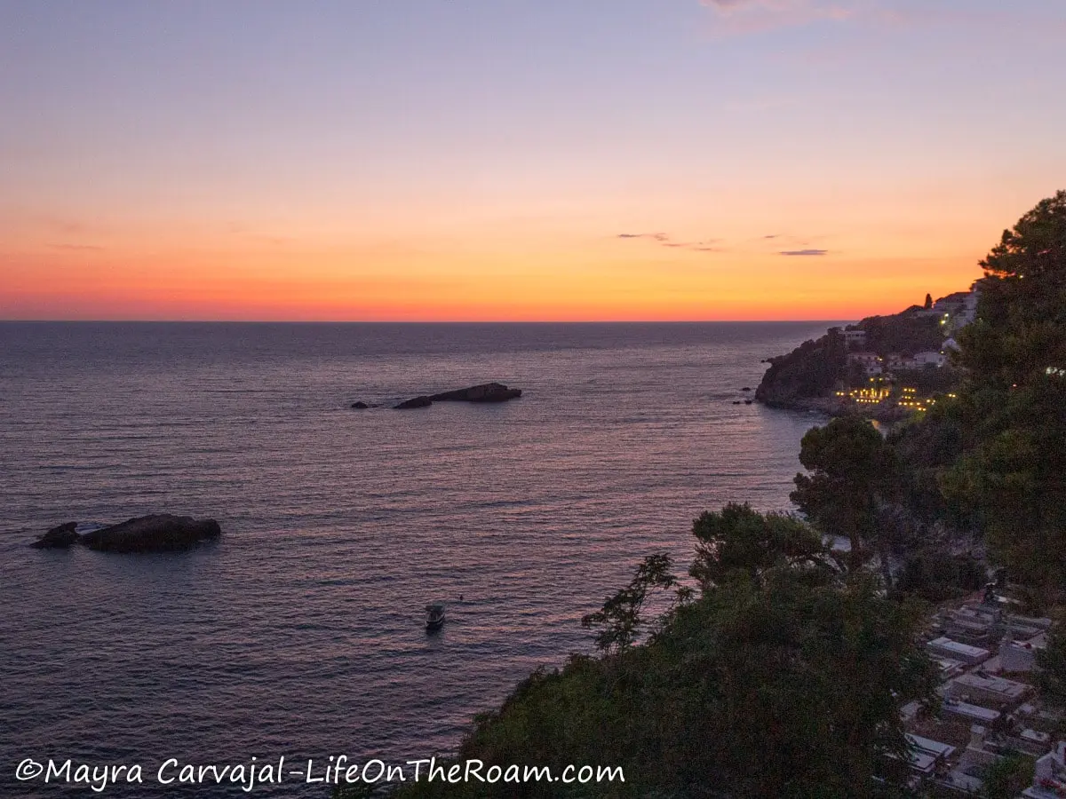 Sunset view from a high point with a coastline with a cemetery and some building, and big rocks in the water