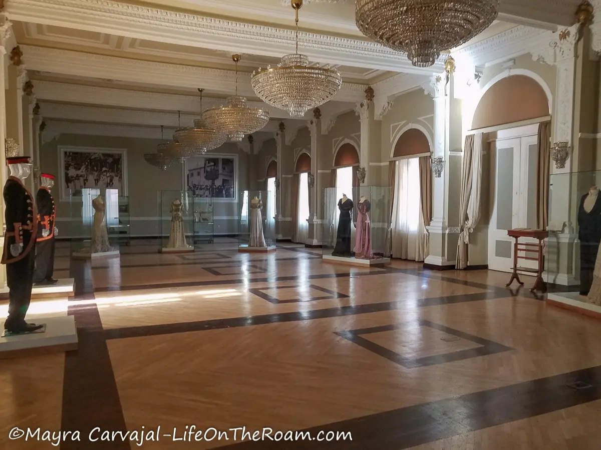 A spacious hall with wood floors and large crystal chandeliers with glass cases exhibiting fine garments