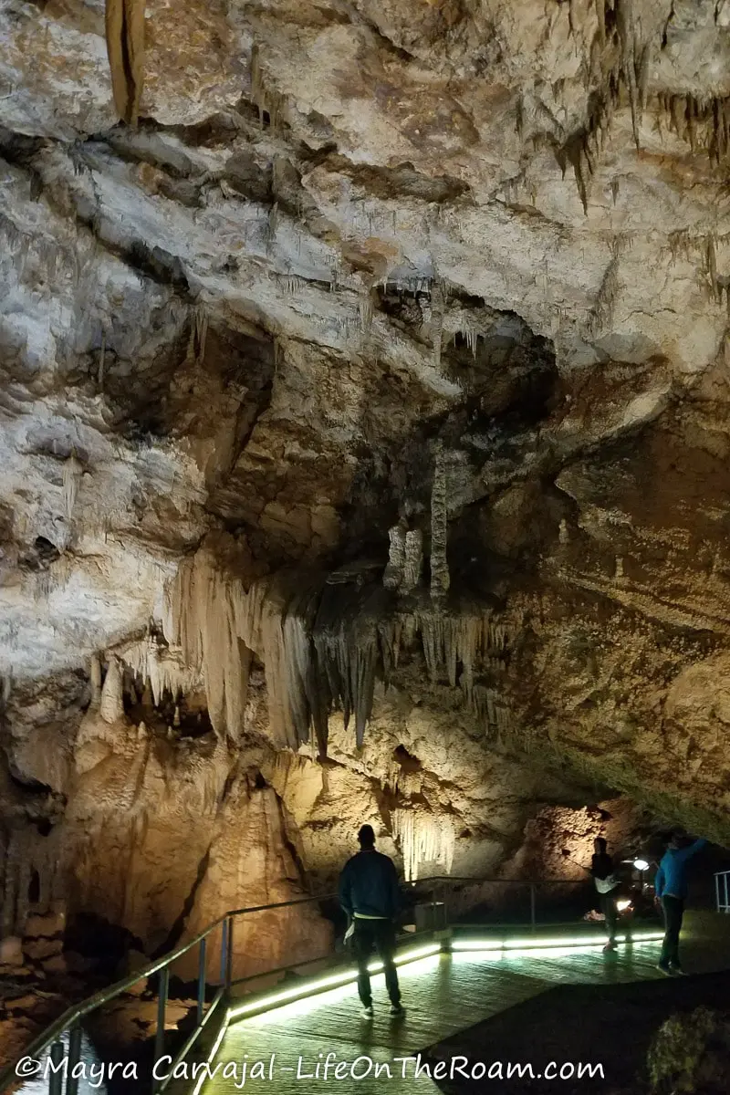 People walking on an illuminated platform inside a tall and wide cave with big stalactites.