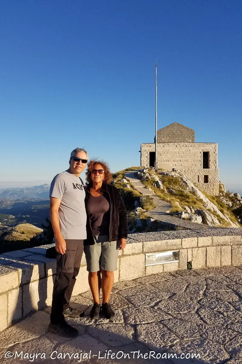 A couple standing on a round lookout overlooking the mountains, with a square building at the back