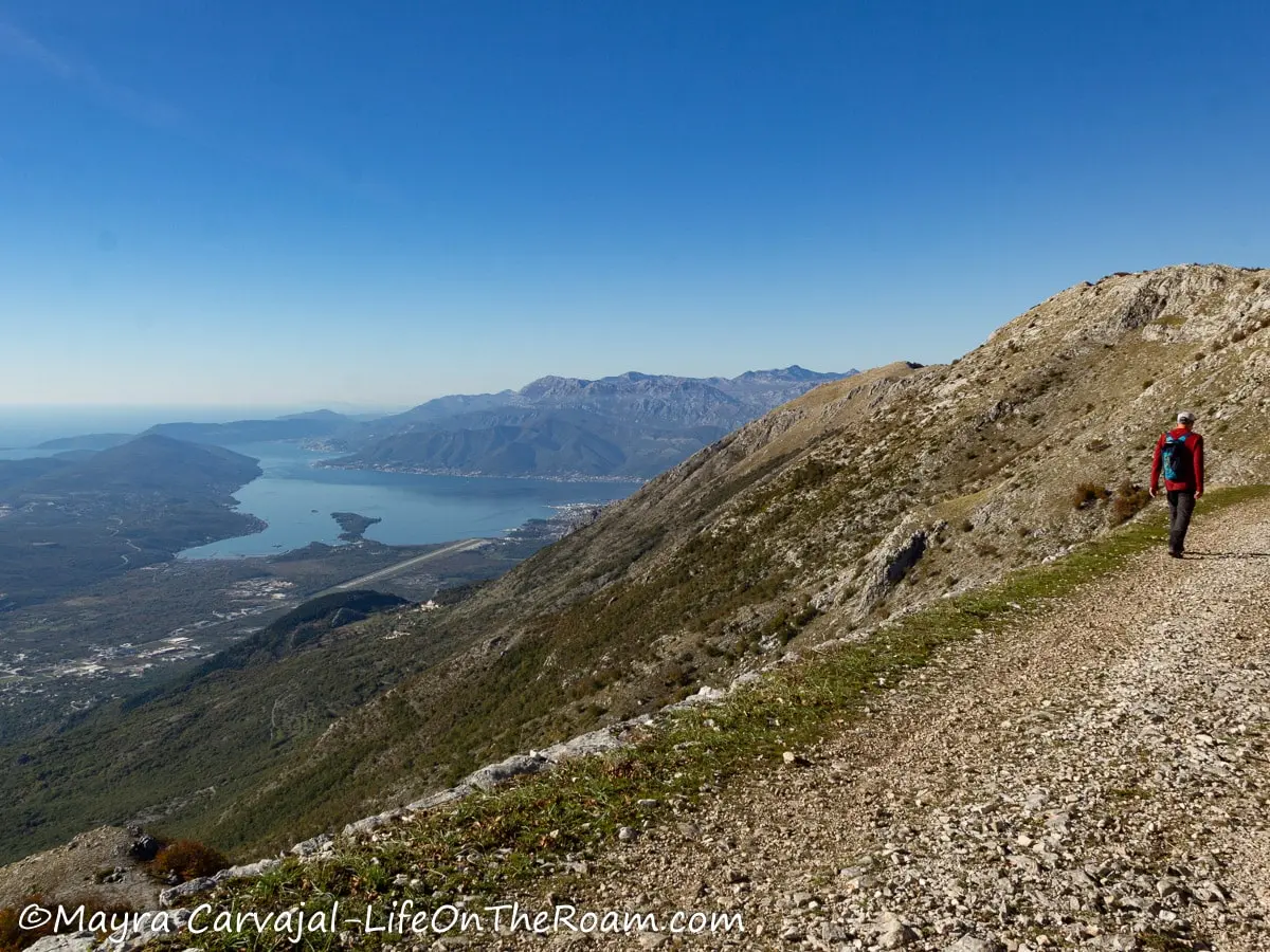 A hiker walking at the edge of a mountain trail with a view of a bay on the left