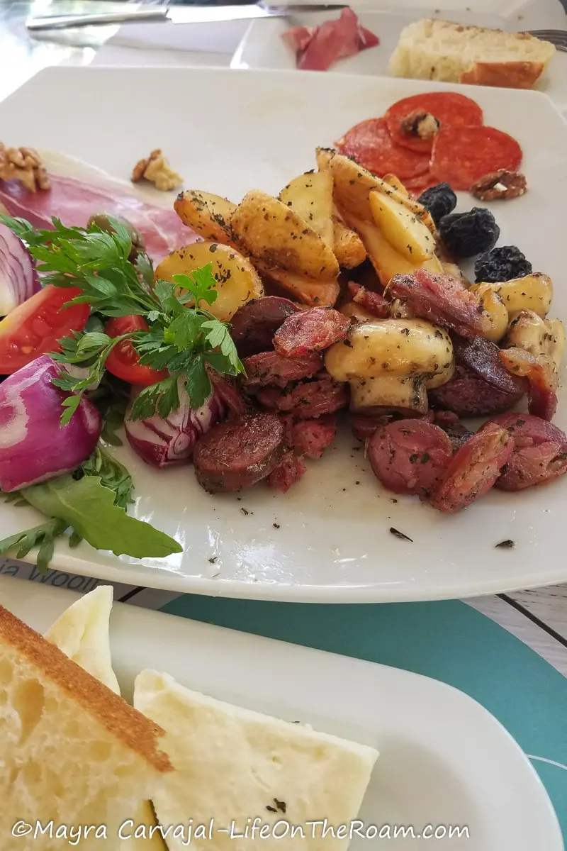 A dish with potato wedges, mushrooms, olives, sausages and a plate with bread and white cheese