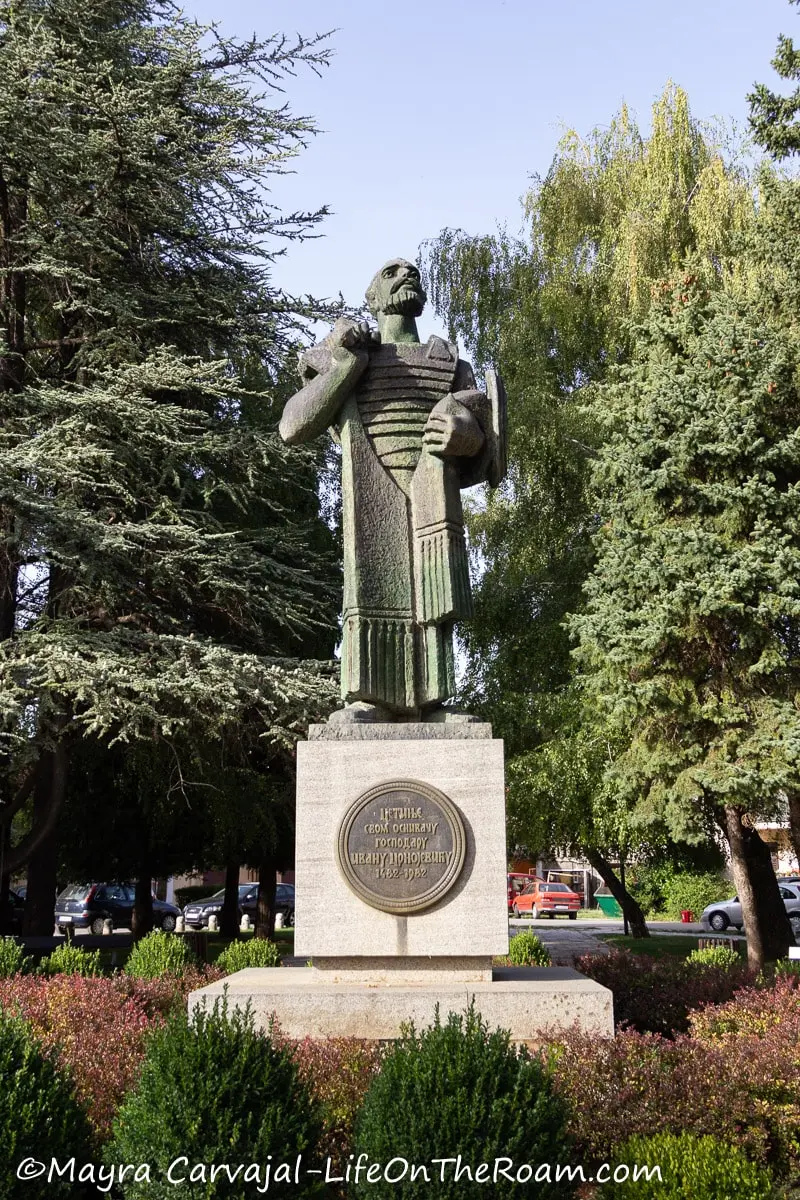 A bronze statue of a man on a square stone base in a park with tall trees