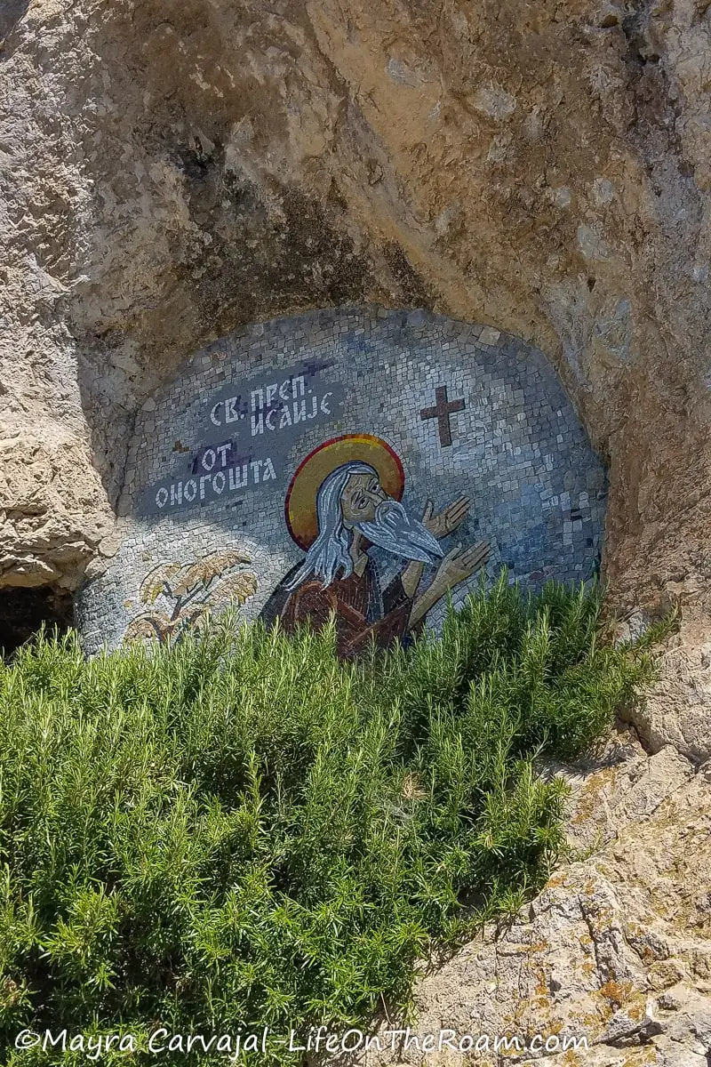 A colourful mosaic on a cliff depicting a saint, with vegetation in the foreground