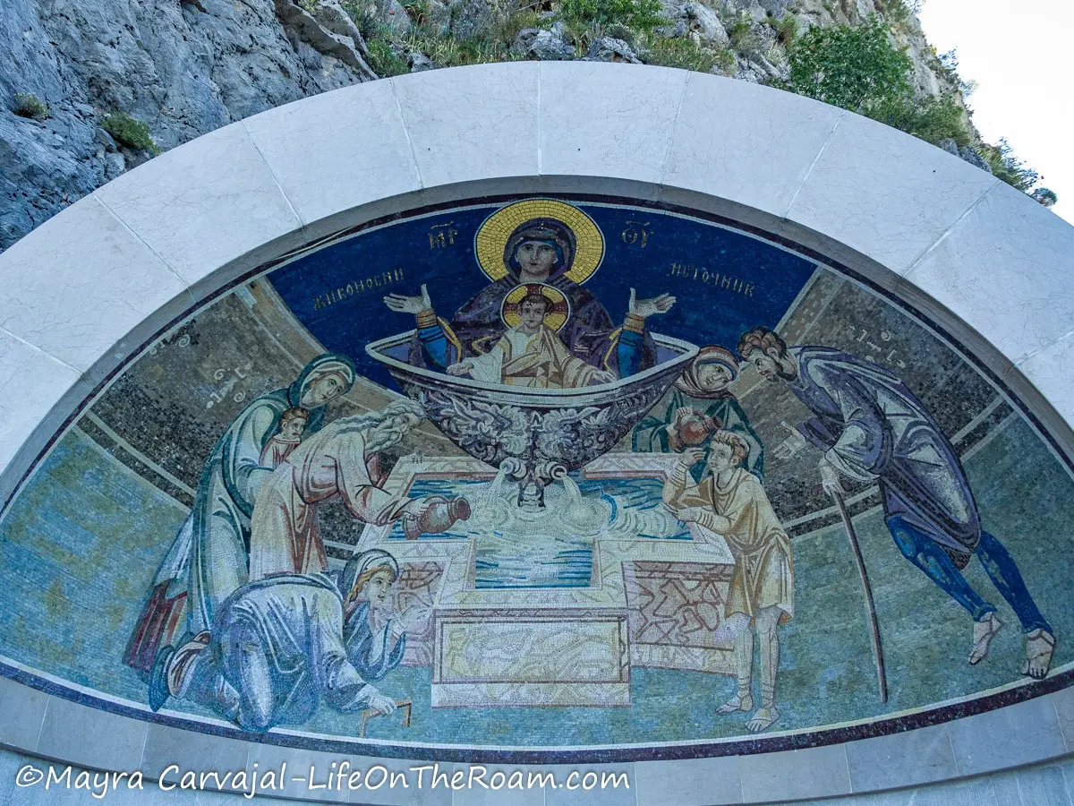 A mosaic under a dome depicting Virgin Mary with Jesus in a fountain that spills in another fountain in the shape of a cross with people around