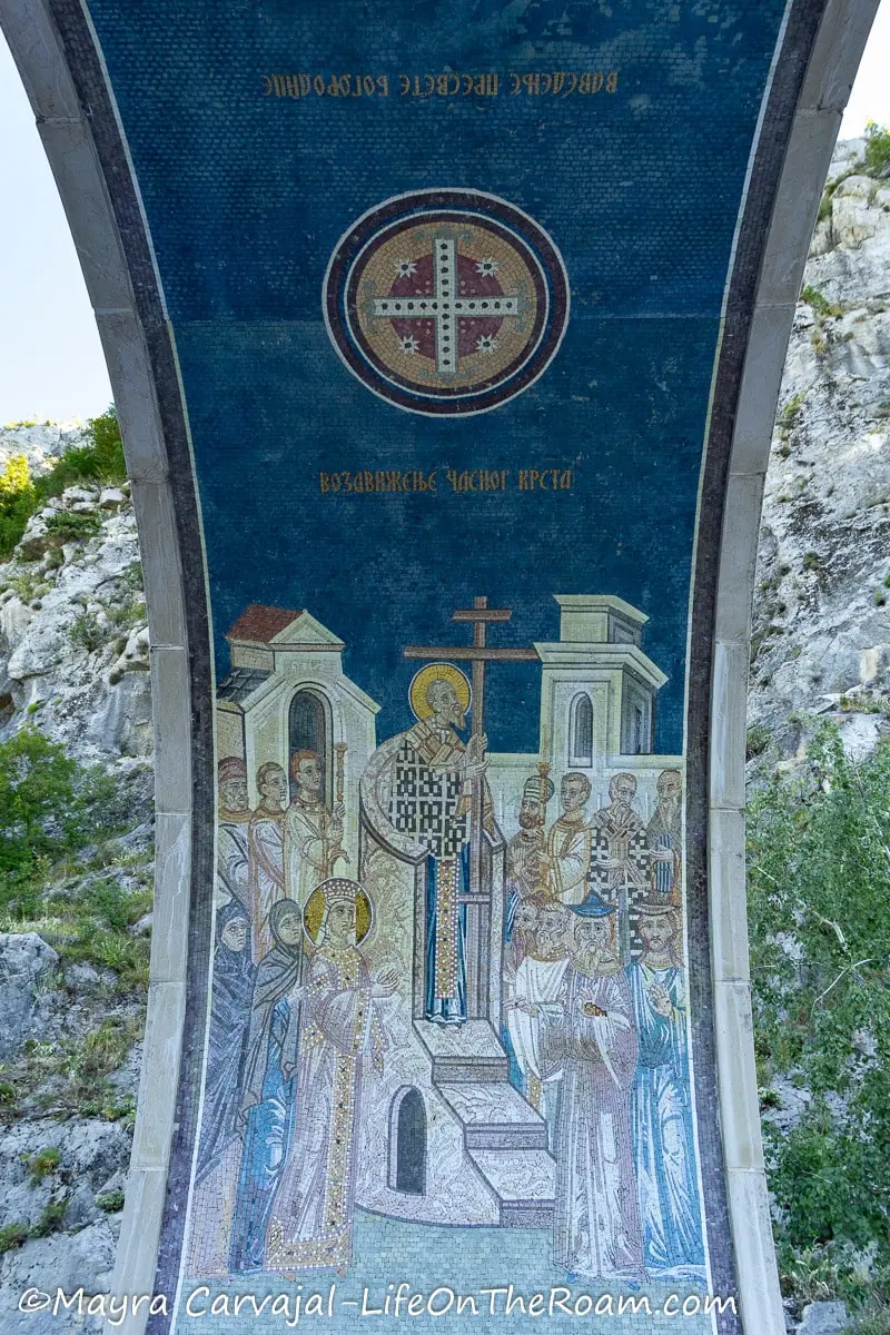 A mosaic under and arch with blue colours depicting a saint with a cross in front of people.