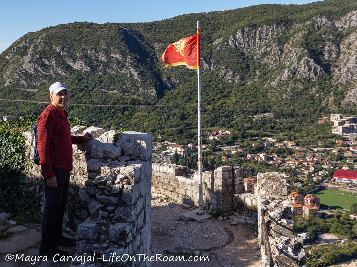 A man standing on a viewpoint with mountain views in the background and the flag of Montenegro