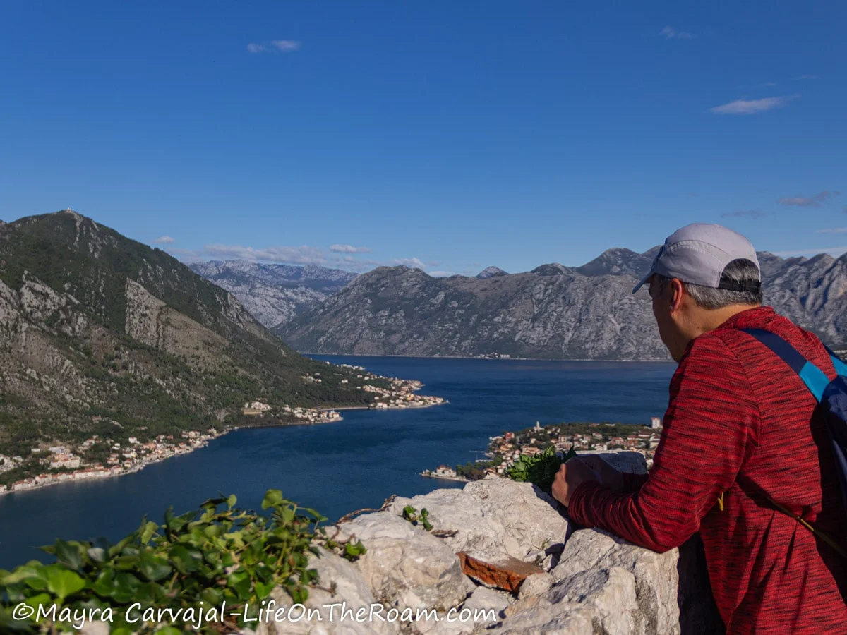 A man at a viewpoint overlooking mountain ridges and a bay with small towns along the shore