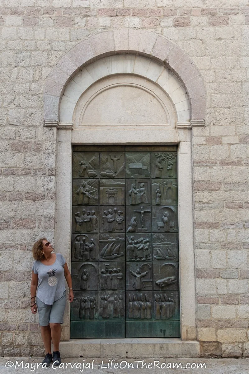 Mayra standing next to a tall bronze double door with 24 panels depicting scenes