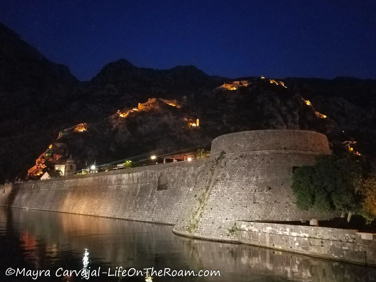 A river along a wall surrounding a fortified town, with night illumination
