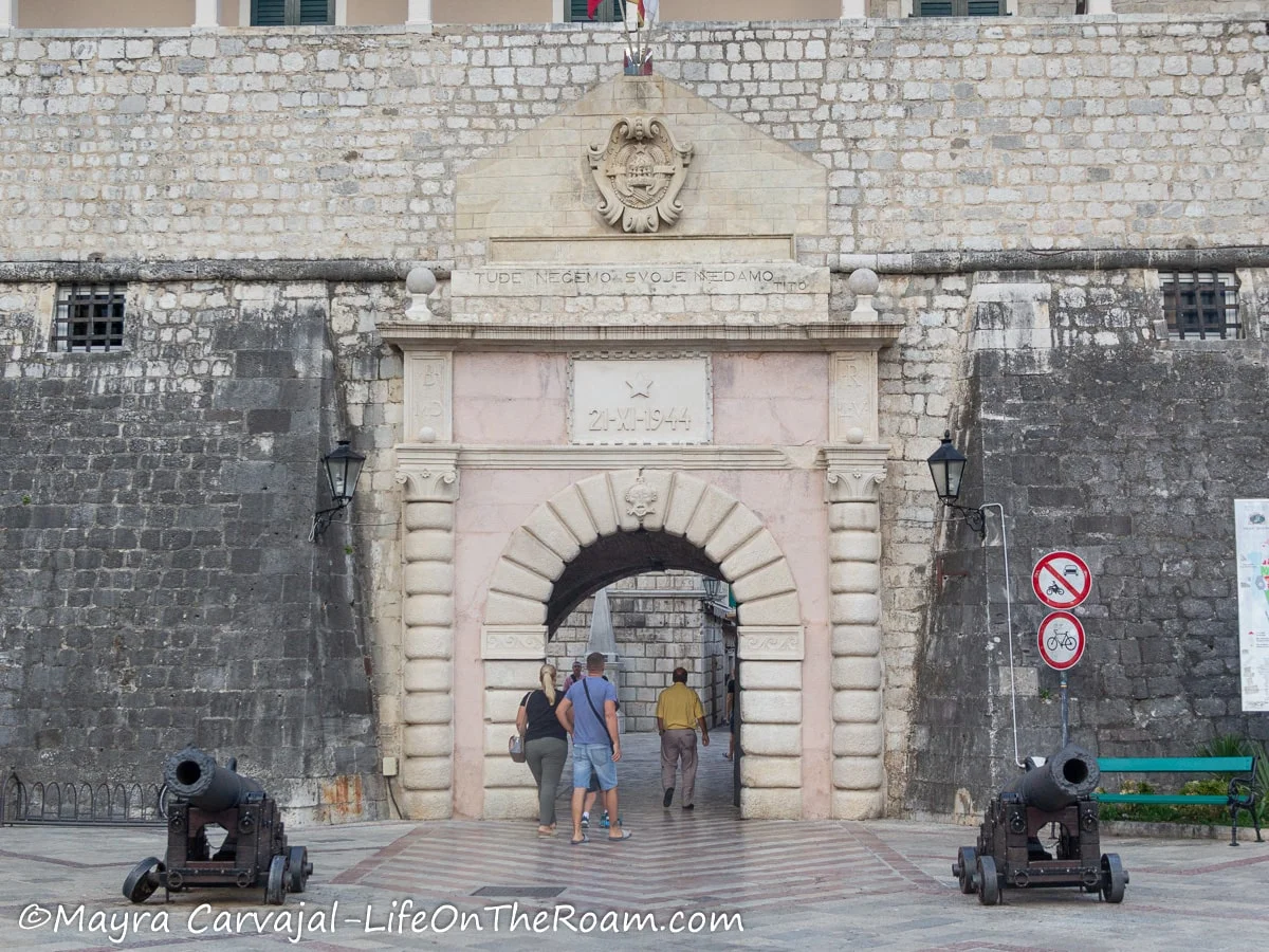 A wide arched gate into a walled town with inscription above and two cannons flanking the entrance