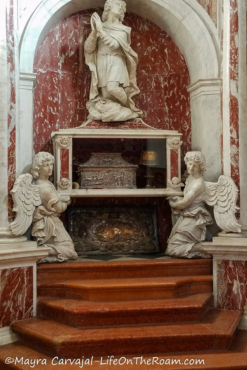 A marble sculpture with two angels holding a case containing a silver casket and a figure in prayer above