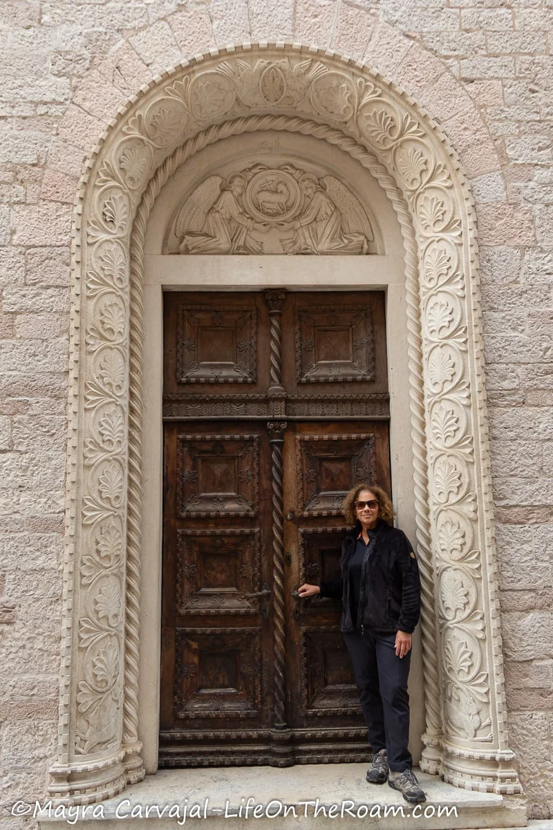 Mayra standing next to a tall wooden double door surrounded by an elaborate trim