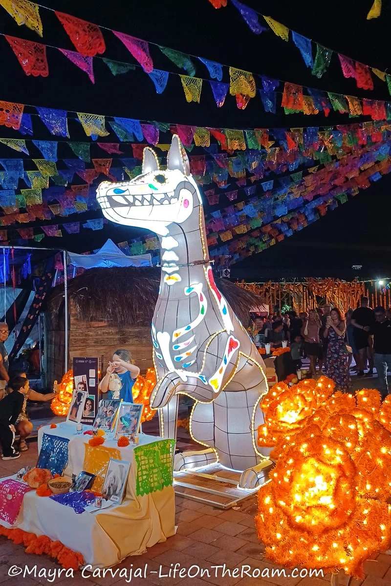 An illuminated big prop of a dog behind a Day of the Dead altar installed in a public square with perforated paper