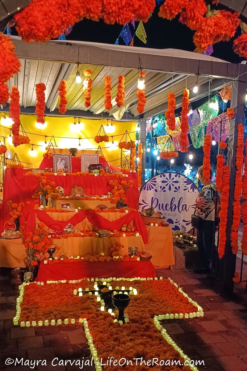 A Day of the Dead altar with an orange tapestry on the floor and a cross made with candles and the sign saying Puebla