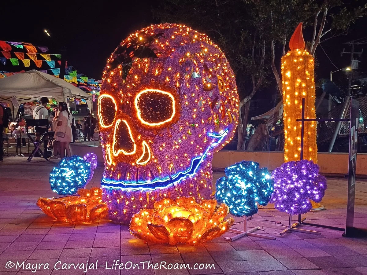 Giant illuminated figures of a skull, marigold flowers and a candle in a public square at night