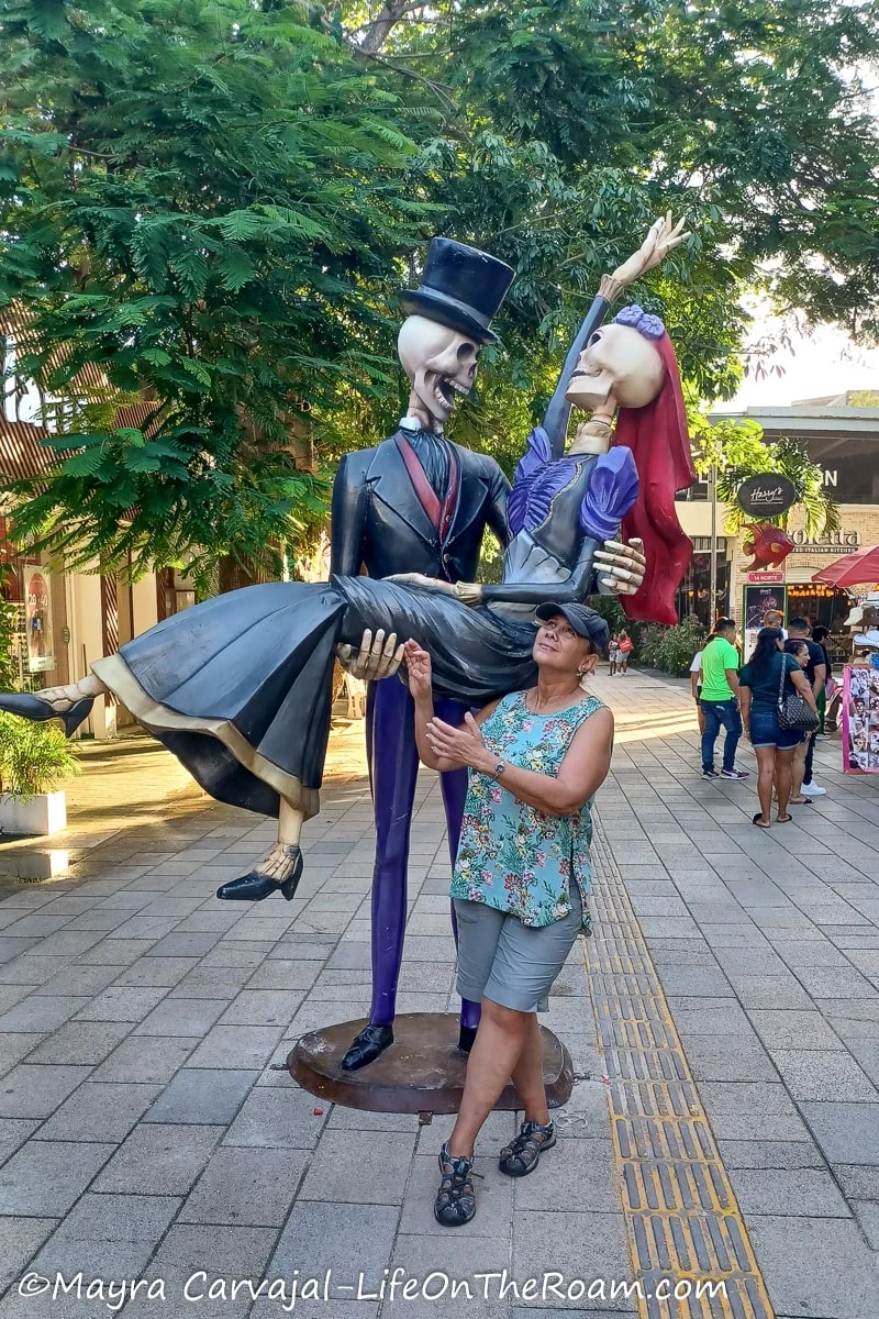 Mayra posing next to a giant decorated skeleton depicting a suited man holding a skeleton lady with a dress in a pedestrian street