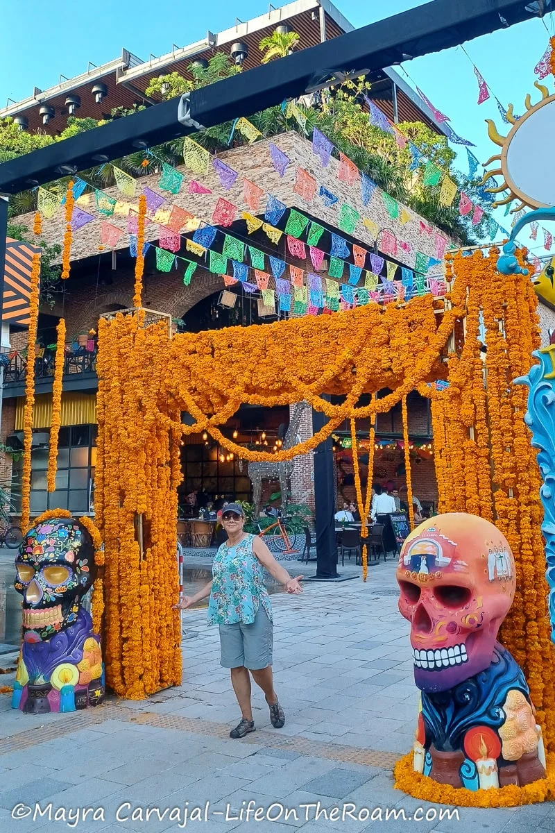 Mayra standing under a decorated arch with fake orange flowers and two huge colourful decorated skulls on each side