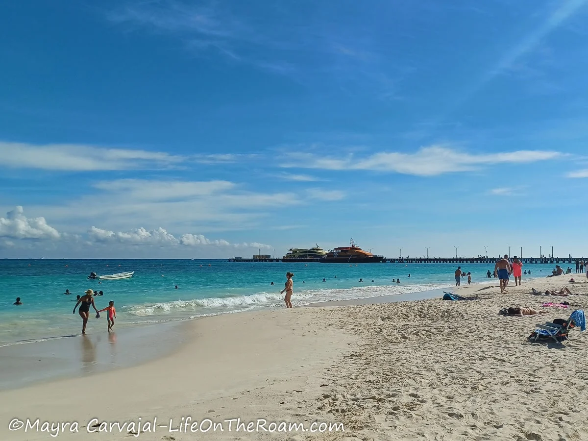 A beach with a blue sea and a wide stretch of sand, with a dock in the background