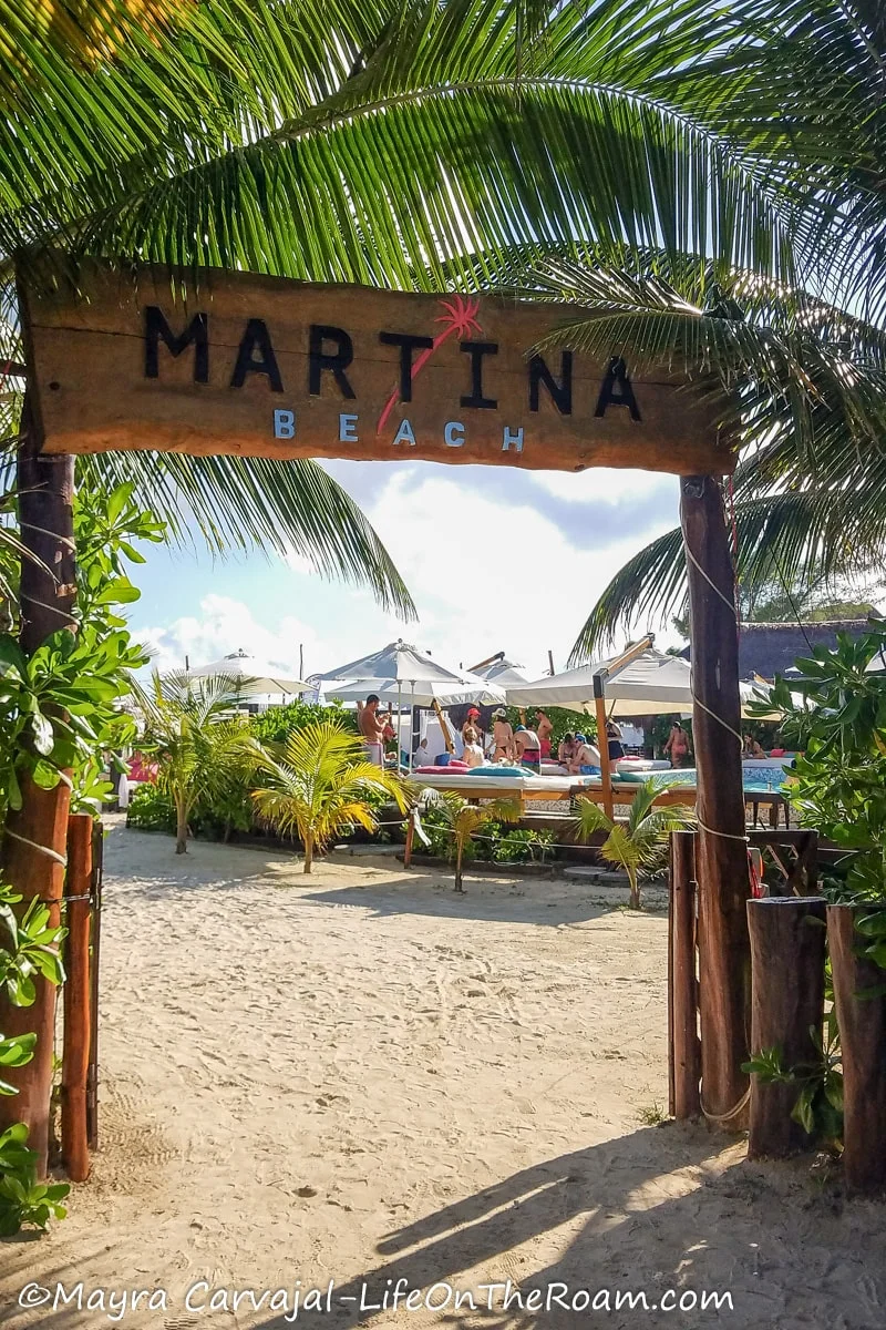 A sign written on a wood board marking the entrance of a beach club reading "Martina Beach"