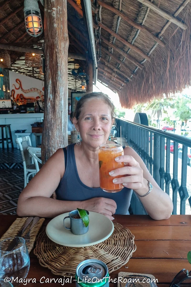 Mayra holding a cocktail glass at a restaurant with a thatch roof