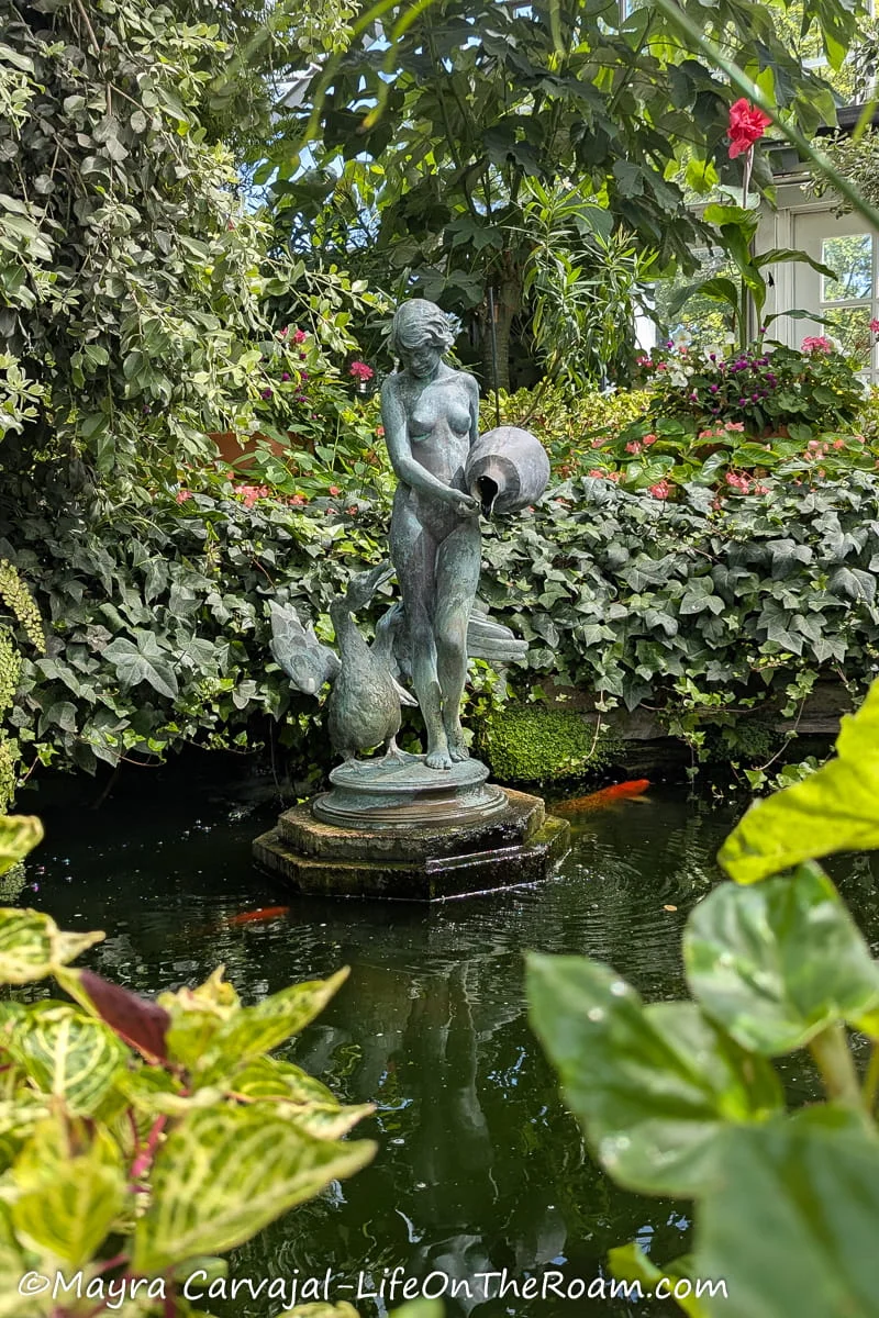 A statue of a woman and a swan on a pond surrounded by plants and with koi fish