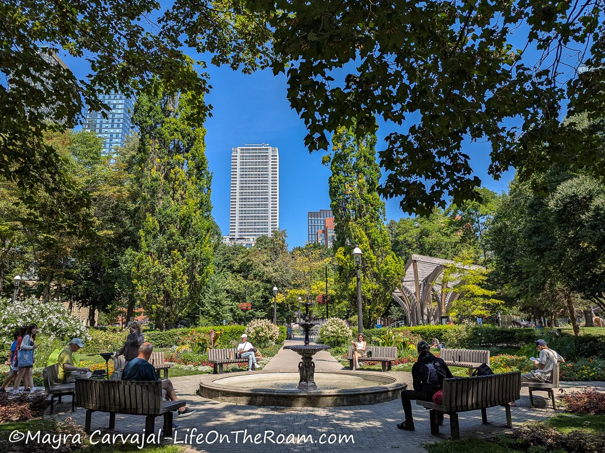 An urban park with an artistic fountain and benches in a circular arrangement and view of skyscrapers in the distances