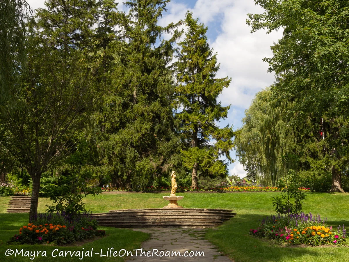A fountain with a statue at the end of a short promenade with trees and flowers